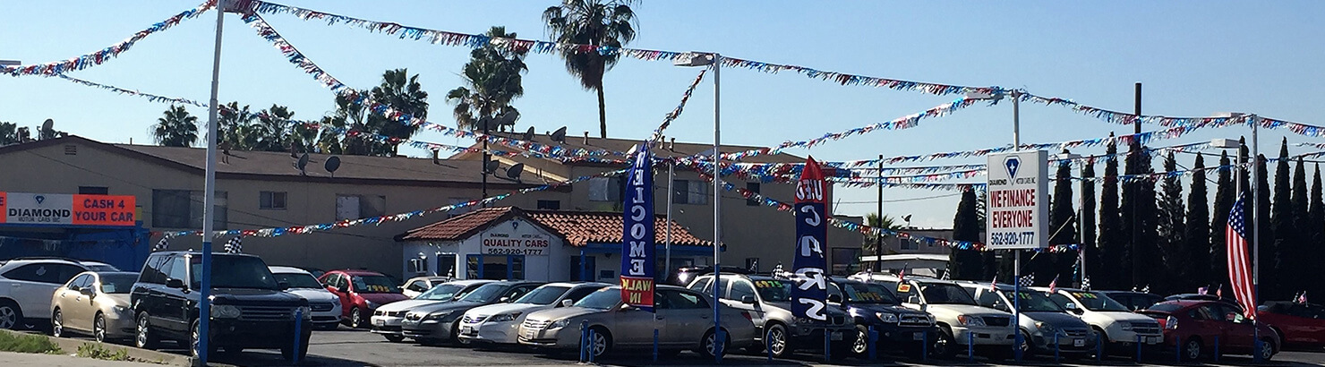 Front of Diamond Motorcars with shiny vehicles for sale, palm trees and flags